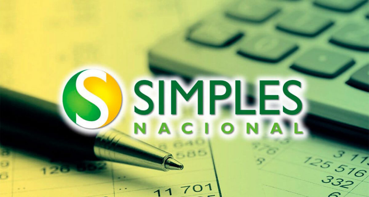 O fundo da imagem é composto por uma caneta, uma calculador e folhas contendo cálculo. No primeiro plano, está a logo do "Simples Nacional".