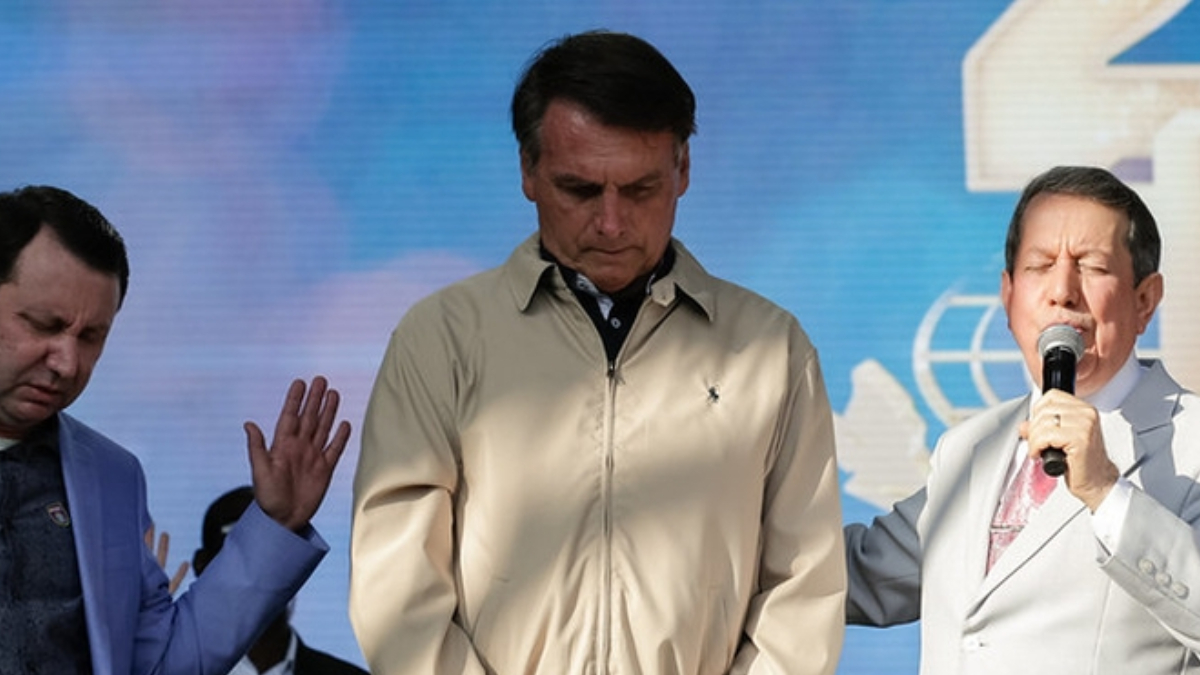A imagem mostra o Presidente Jair Messias Bolsonaro entre Davi Soares e R.R. Soares, pastor e missionário da Igreja Internacional da Graça de Deus. Os três estão de olhos fechado e orando.