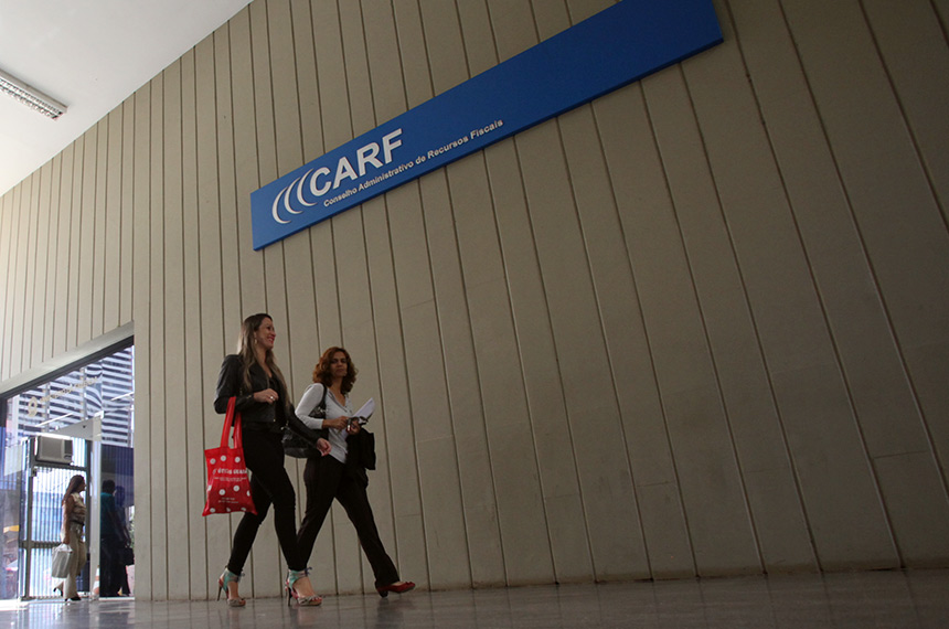 A imagem mostra duas mulheres caminhando de frente ao CARF (Conselho Administrativo de Recursos Fiscais). No topo superior da imagem uma placa indica o CARF.