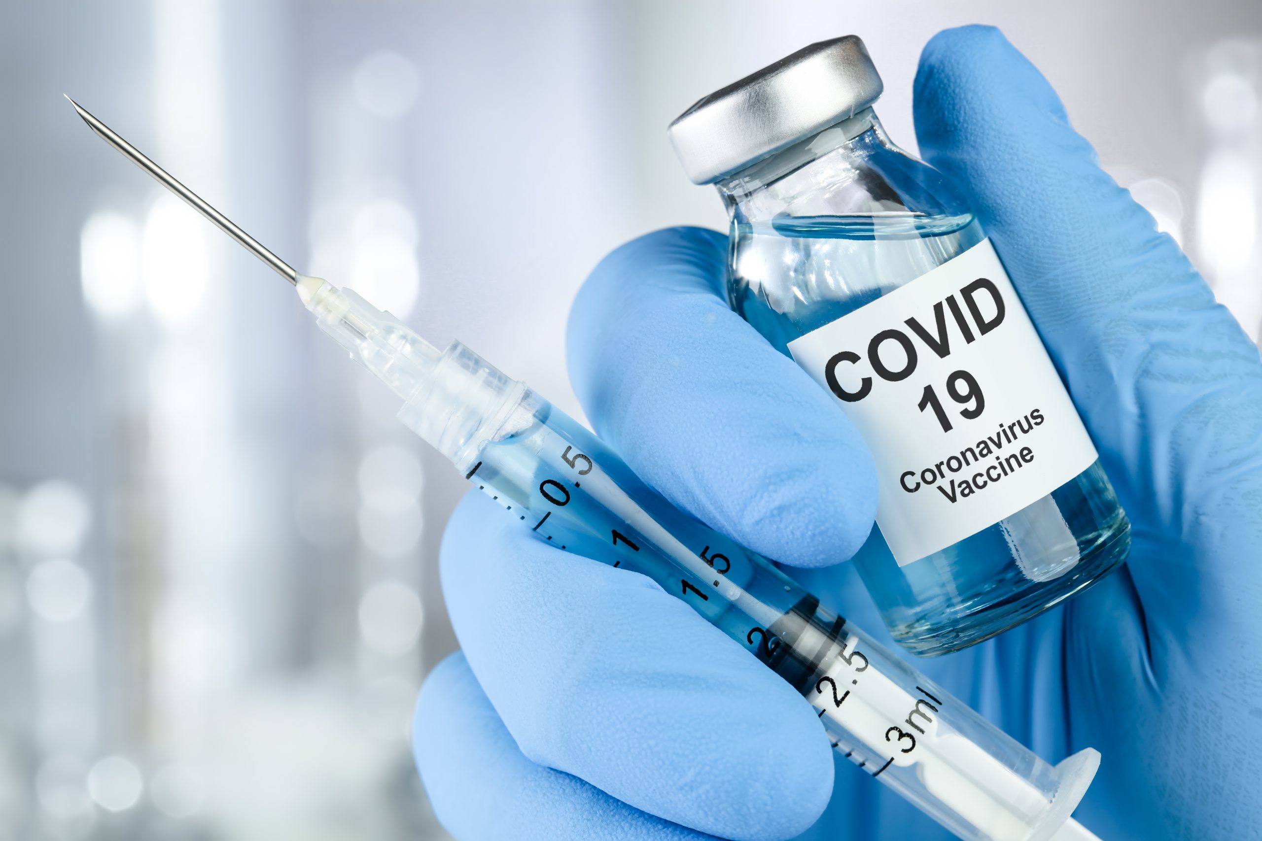 A imagem mostra uma mão vestida com luva azul, segurando um recipiente com a vacina contra o COVID-19 e uma seringa cheia do líquido.