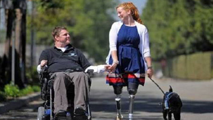 A imagem mostra um homem cadeirante de mãos dadas com uma mulher com prótese nas duas pernas. A mulher segura um cachorro preto pela coleira.