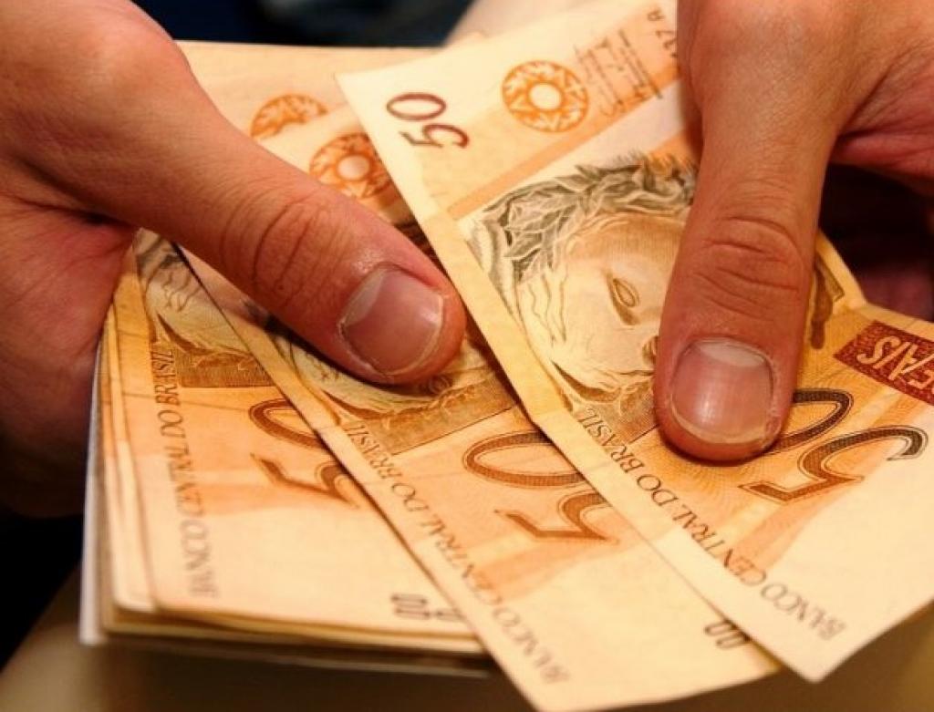 Duas mãos segurando diversas notas de R$ 50,00 (cinquenta reais)